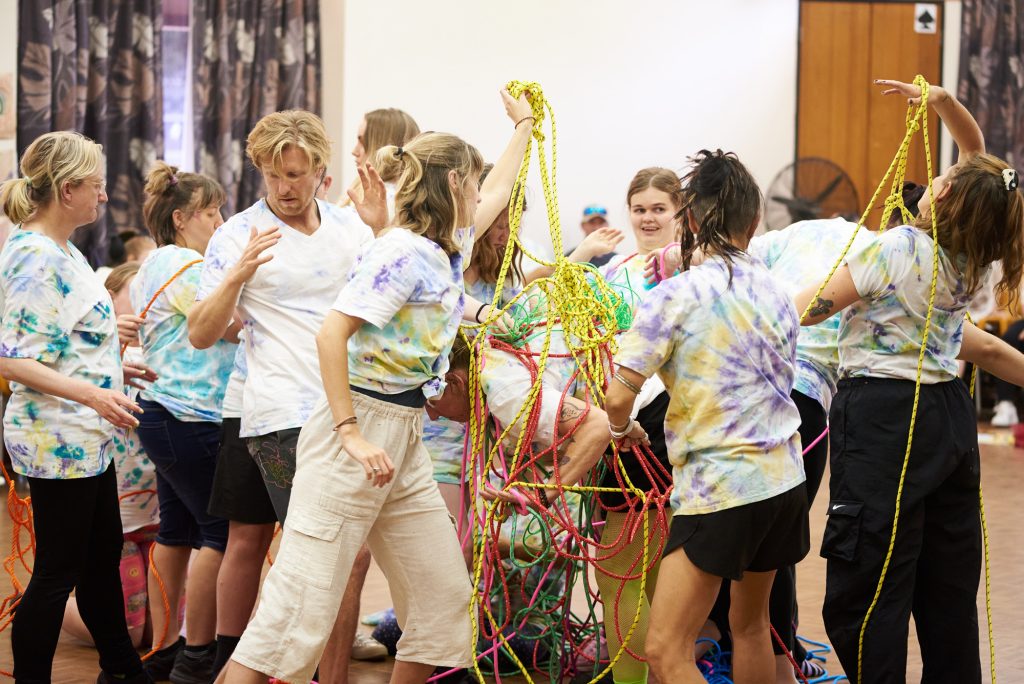 A large group of performers in tie-dyed t-shirts are entwined together with a mess of brightly coloured ropes.
