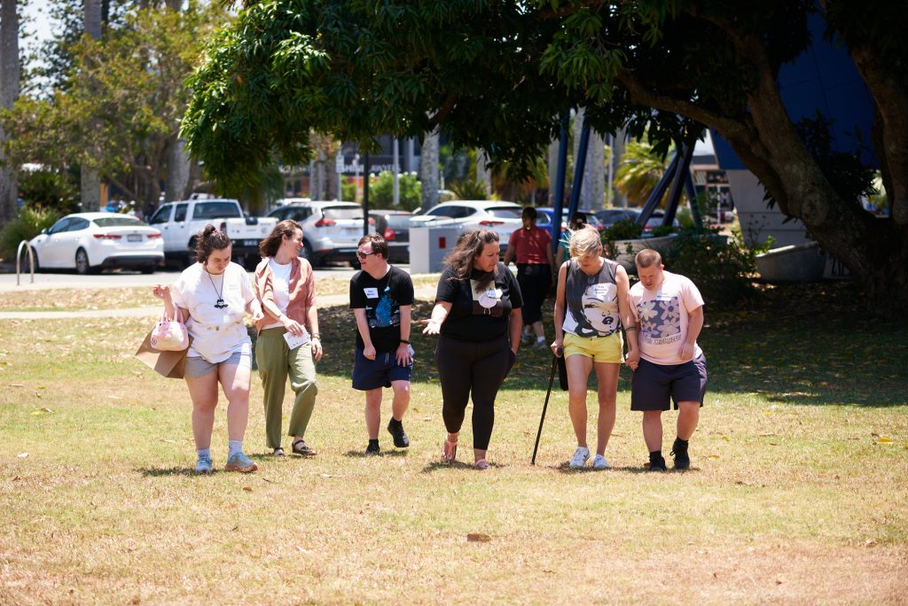 A candid photo of a group of six people walking in a line across a grassed area toward the camera, one person is using a walking cane.